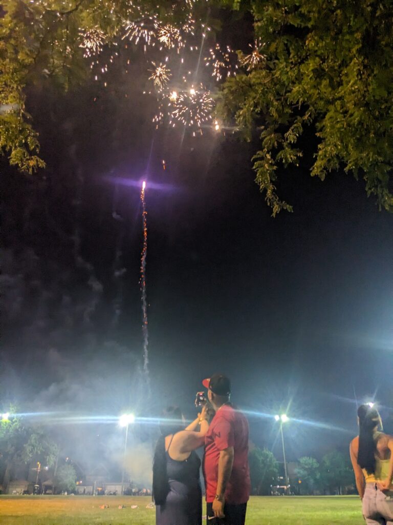 A man and woman stand beside each other in an open field under bright lights, filming a fireworks display for which they are responsible.